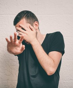 Een man staat voor een witte muur, hij maakt een angstige indruk, verstopt zich achter zijn hand, spiekt tussen zijn vingers door en steekt zijn andere hand afwerend voor zich uit.